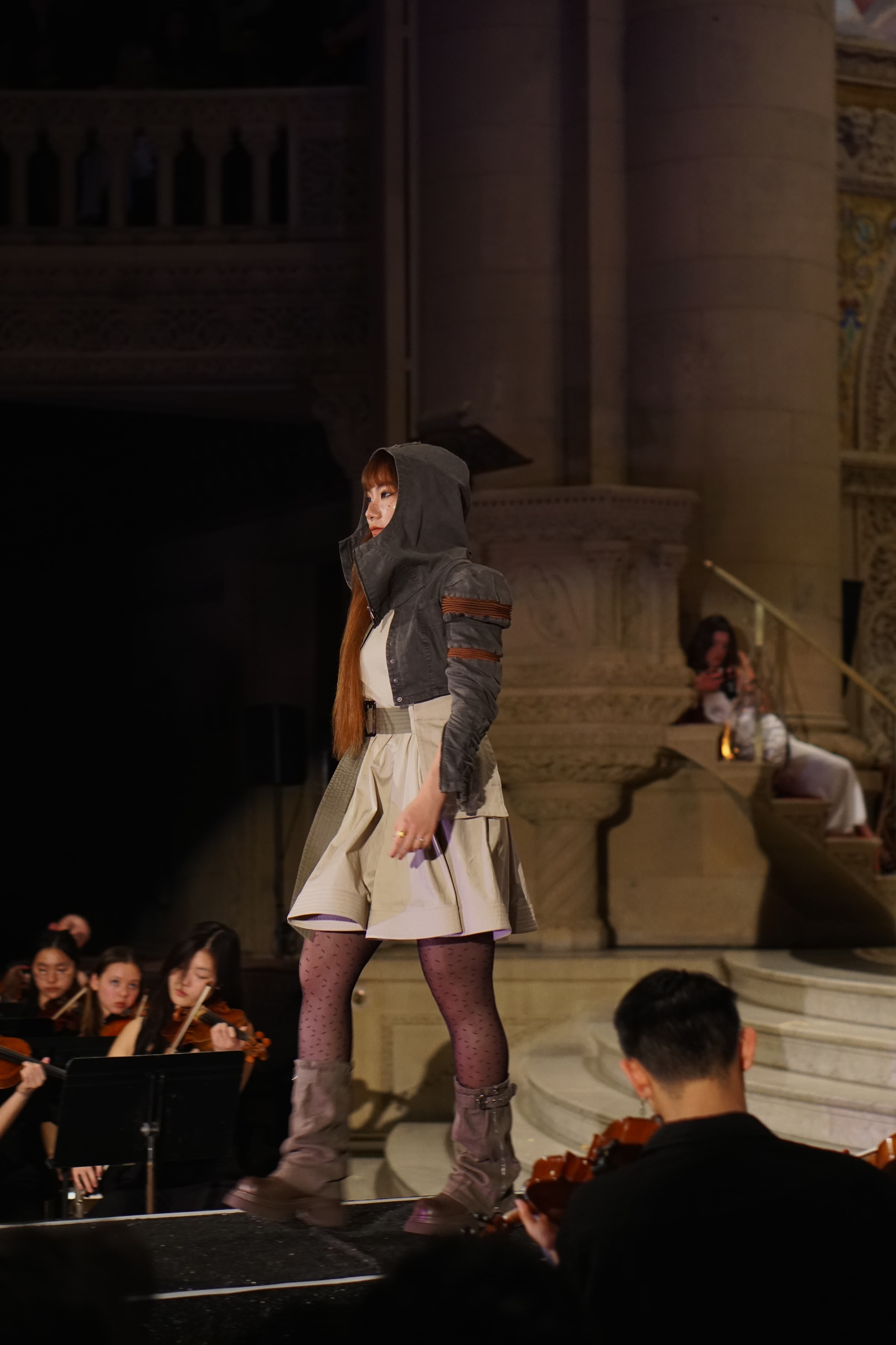 Model in hooded jacket and pleated skirt with orchestra in background
